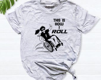 Puede incluir: Una camiseta gris claro de manga corta con un gráfico negro de una persona en silla de ruedas haciendo un gesto de pulgar hacia arriba. El texto dice THIS IS HOW I ROLL. La camiseta tiene cuello redondo y un ajuste casual con un logo.
