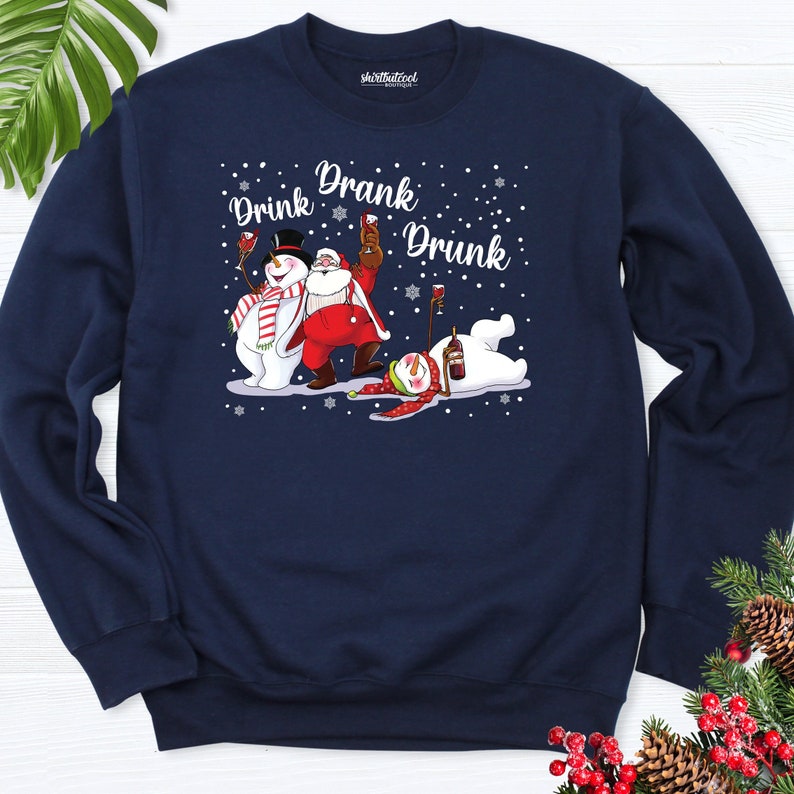 May include: Navy blue sweatshirt with a festive graphic featuring a snowman, Santa Claus, and a snowman, all holding wine glasses. The text "Drink Drank Drunk" is displayed above the characters in a white font. Snowflakes are scattered around the characters.