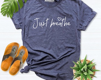 May include: A heather navy blue short-sleeve t-shirt with a crew neckline. The front features the white text "Just breathe" in a cursive script font, with a small heart icon above the letter "r". The brand name "shirtbutcool BOUTIQUE" is printed in white near the collar.