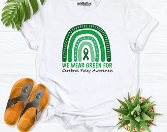 May include: A white short-sleeve t-shirt featuring a green rainbow graphic with a central awareness ribbon. The text reads WE WEAR GREEN FOR Cerebral Palsy Awareness. The shirt includes a small shirtbutcool boutique logo at the neck.