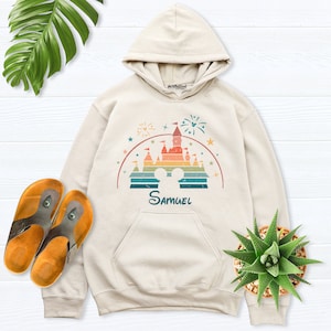 Puede incluir: Una sudadera con capucha beige con un gr&aacute;fico de estilo vintage de un castillo con fuegos artificiales y el nombre "Samuel" impreso debajo. El gr&aacute;fico est&aacute; en colores arco&iacute;ris con un aspecto desgastado.