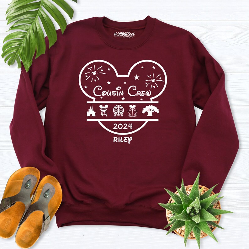 May include: A maroon sweatshirt with a white graphic that reads "Cousin Crew 2024 Riley" in a Mickey Mouse ear design. The graphic includes fireworks, stars, and various park icons. The sweatshirt is displayed with sandals and a small succulent.