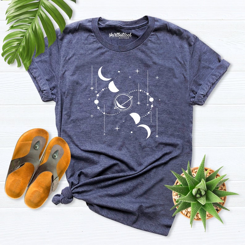 May include: A navy blue t-shirt with a white graphic of a celestial design featuring moons, stars, and a planet. The text "shirtbutcool" is printed on the shirt.