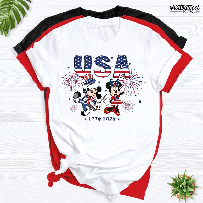 Pode incluir: Uma t-shirt branca com um gr&aacute;fico do Mickey e da Minnie vestidos com roupas patri&oacute;ticas com o tema da bandeira americana. Acima, o texto USA est&aacute; escrito com um padr&atilde;o de bandeira e, abaixo, o texto 1776-2026 &eacute; exibido com fogos de artif&iacute;cio.