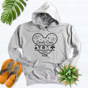 May include: A light gray hooded sweatshirt with a black graphic design. The design features a Mickey Mouse head shape with the words "Cousin Crew 2024 Riley" and various icons. The sweatshirt has a front pocket and a drawstring hood.