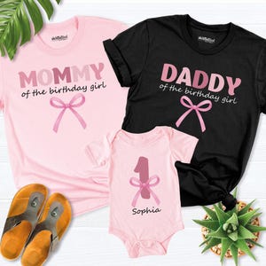 May include: Pink t-shirt with "MOMMY of the birthday girl" text and a pink bow graphic. A black t-shirt with "DADDY of the birthday girl" text and a pink bow graphic. A pink baby onesie with a number 1 and a pink bow graphic, with the name "Sophia".