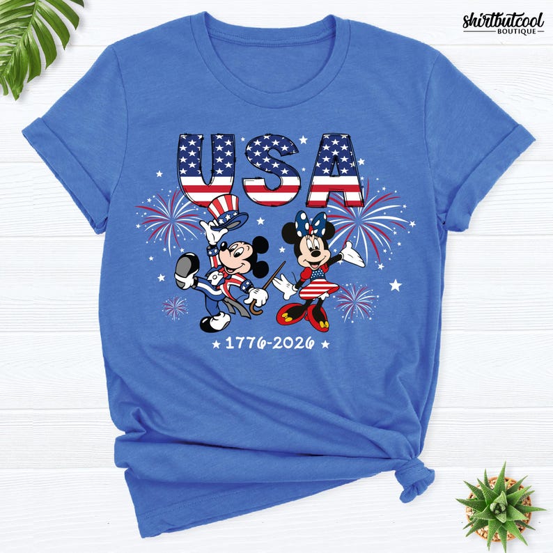 Pode incluir: Uma t-shirt azul brilhante com um gr&aacute;fico do Mickey e da Minnie vestidos com roupas patri&oacute;ticas vermelhas, brancas e azuis. Acima, o texto USA est&aacute; impresso com um padr&atilde;o de bandeira, com fogos de artif&iacute;cio e o texto 1776-2026 abaixo.
