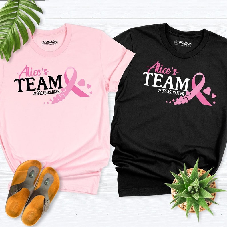 May include: Two t-shirts with the text "Alice's Team #BreastCancer" and a pink ribbon with hearts. The shirts are pink and black.