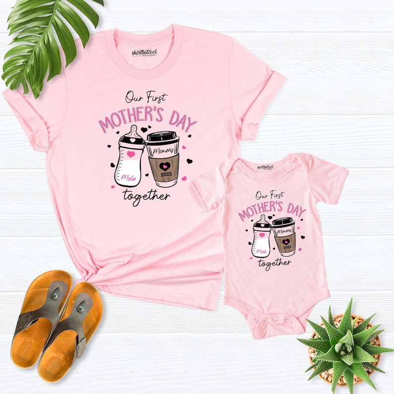 May include: Pink t-shirt and bodysuit set with a graphic of a baby bottle and a coffee cup. The text reads "Our First Mother's Day Together".