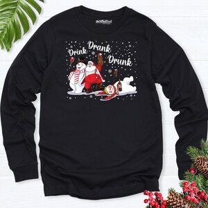 May include: Black long-sleeve shirt with a graphic of a snowman, Santa Claus, and a polar bear, all appearing intoxicated. The words "Drink, Drank, Drunk" are above the characters. Snowflakes are scattered around the characters.