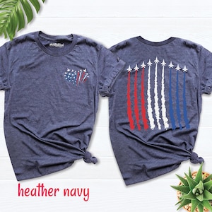 May include: A heather navy t-shirt featuring a small heart-shaped graphic on the front left chest composed of jets and red, white, and blue smoke trails. The back displays a row of eight jets flying upward with long red, white, and blue smoke trails.