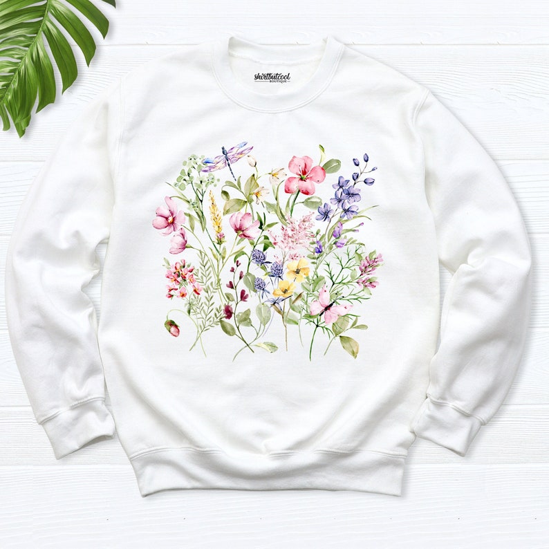 K&ouml;nnte beinhalten: Wei&szlig;er Pullover mit einem Blumendruck mit rosa, lila, gelben und gr&uuml;nen Aquarellblumen und einer Libelle.