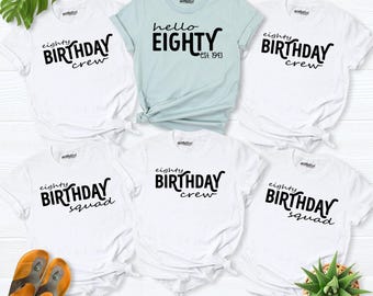 May include: Five white t-shirts and one light blue t-shirt with black text. The white shirts feature the text 'eighty BIRTHDAY crew' or 'eighty BIRTHDAY squad'. The light blue shirt features the text 'hello EIGHTY est. 1943'.