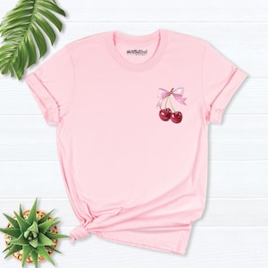 May include: Pink t-shirt with a graphic of two cherries with a pink bow on a white background.