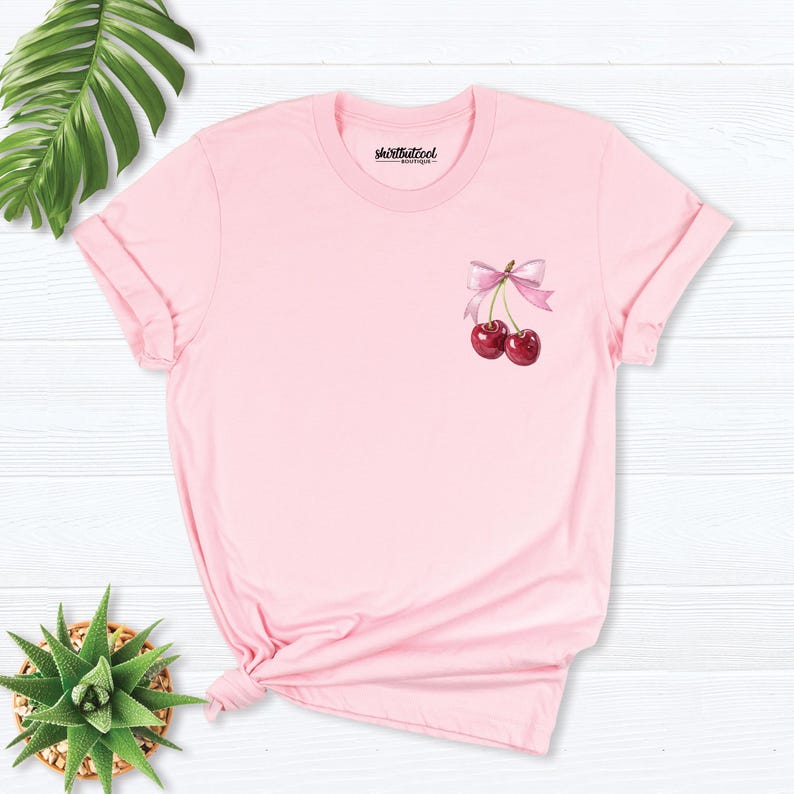 May include: Pink t-shirt with a graphic of two cherries with a pink bow on a white background.