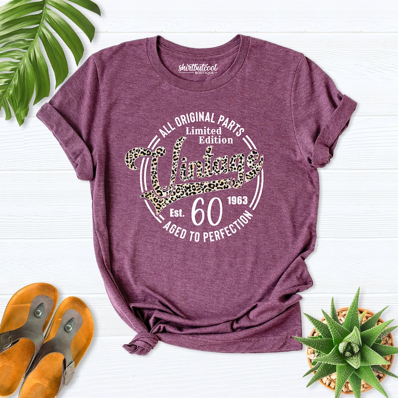 K&ouml;nnte beinhalten: Ein lila T-Shirt mit einem wei&szlig;en Kreis-Motiv, auf dem "All Original Parts Limited Edition Vintage Est. 60 1963 Aged to Perfection" steht. Das Motiv hat ein Leopardenmuster.