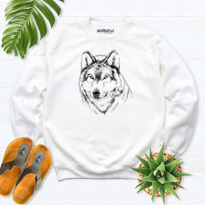 Pu&ograve; includere: Felpa bianca con un'illustrazione in bianco e nero di una testa di lupo. Il testo "shirtbutcool" &egrave; stampato sulla felpa.