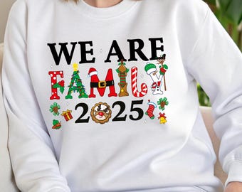 May include: A white long-sleeve sweatshirt featuring the black text WE ARE FAMILY 2025. The letters in FAMILY are decorated with Christmas motifs including a tree, Santa belt, candy cane, and snowman. Small holiday icons surround the text.