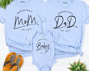 May include: Three light blue garments including two short-sleeve t-shirts and one baby bodysuit. Each features black text reading Emerson Family, Est. 2025, and the words Mom, Dad, or Baby in a script font. The shirts have a small logo at the neck.