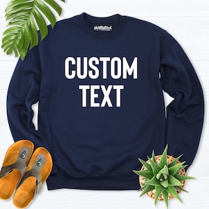 Pode incluir: Uma camisola de manga comprida azul-marinho com gola redonda, apresentando o texto branco CUSTOM TEXT impresso numa fonte sem serifa em negrito no peito. A peça inclui uma pequena etiqueta interior com SHIRTBUTCOOL BOUTIQUE.