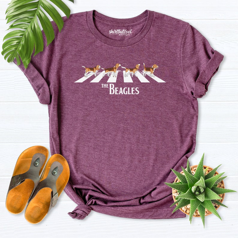 Puede incluir: Una camiseta morada con un gr&aacute;fico de cinco perros beagle caminando por un paso de cebra. El texto "The Beagles" est&aacute; impreso debajo de los perros.