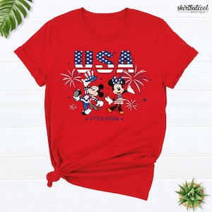 Pode incluir: Uma t-shirt vermelha de manga curta com um gr&aacute;fico do Mickey e da Minnie em trajes patri&oacute;ticos. Acima, o texto USA est&aacute; impresso com um padr&atilde;o de estrelas e riscas. Fogo de artif&iacute;cio e o texto 1776-2026 tamb&eacute;m s&atilde;o vis&iacute;veis na frente.