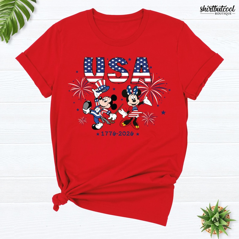 Pode incluir: Uma t-shirt vermelha de manga curta com um gr&aacute;fico do Mickey e da Minnie em trajes patri&oacute;ticos. Acima, o texto USA est&aacute; impresso com um padr&atilde;o de estrelas e riscas. Fogo de artif&iacute;cio e o texto 1776-2026 tamb&eacute;m s&atilde;o vis&iacute;veis na frente.