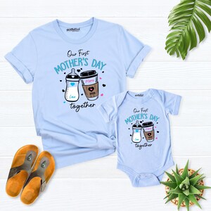 May include: A light blue t-shirt and a light blue bodysuit with the text "Our First Mother's Day Together" and a graphic of a baby bottle and a coffee cup. The coffee cup has the text "Mama 2023" on it.