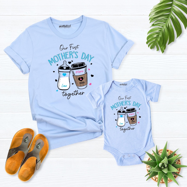 May include: A light blue t-shirt and a light blue bodysuit with the text "Our First Mother's Day Together" and a graphic of a baby bottle and a coffee cup. The coffee cup has the text "Mama 2023" on it.