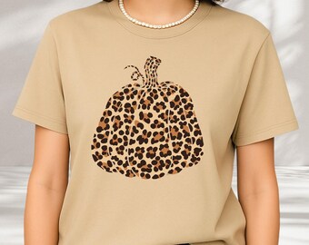 May include: A tan short-sleeve t-shirt featuring a central graphic of a pumpkin with a brown and black leopard print pattern. The shirt has a crew neckline and is worn with a white beaded necklace. The text shirtbutcool boutique is visible.