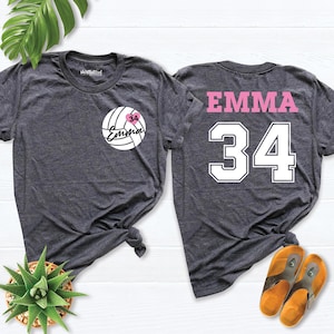 May include: Two dark gray t-shirts. One features a volleyball design with the name "Emma" and the number "34" in white. The other shirt has "Emma" and "34" printed on the back in pink and white. The shirts are displayed on a white surface.