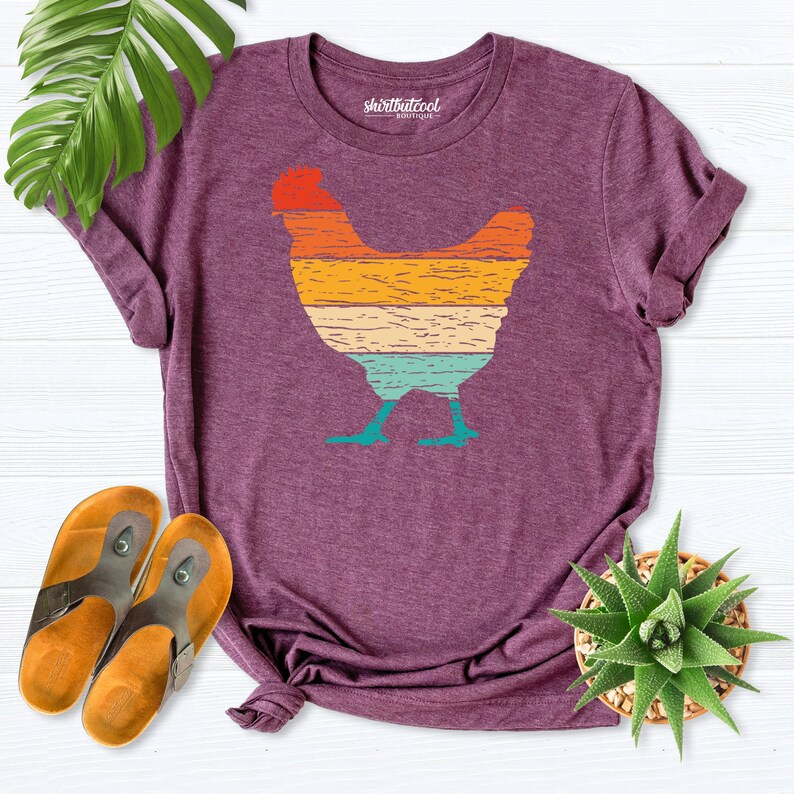 May include: A purple t-shirt with a vintage-style graphic of a chicken. The graphic is made up of horizontal stripes in shades of orange, yellow, green, and blue. The text "shirtbutcool boutique" is printed on the shirt.