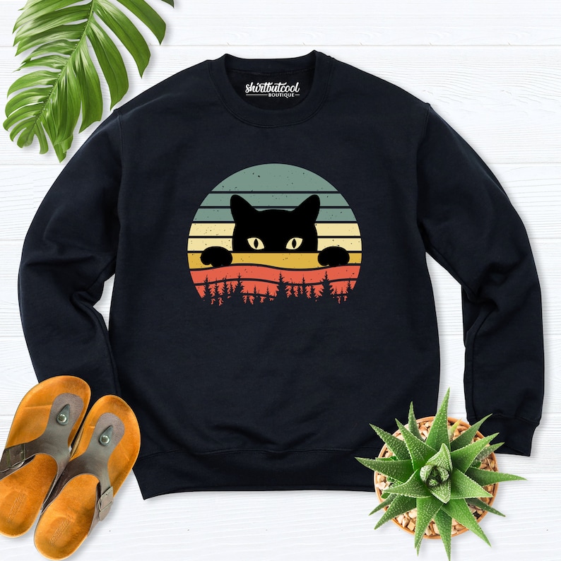 K&ouml;nnte beinhalten: Schwarzes Sweatshirt mit einem Retro-Sonnenuntergang-Design, das eine schwarze Katze zeigt, die &uuml;ber die Baumwipfel schaut. Der Text "shirtbutcool boutique" ist auf dem Sweatshirt aufgedruckt.