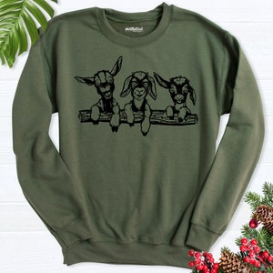 K&ouml;nnte beinhalten: Ein gr&uuml;nes Sweatshirt mit einer schwarzen Grafik von drei jungen Ziegen, die &uuml;ber einen Baumstamm schauen. Die Ziegen haben schwarze Augen und Nasen und wei&szlig;e Markierungen im Gesicht.