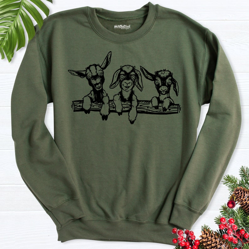 K&ouml;nnte beinhalten: Ein gr&uuml;nes Sweatshirt mit einer schwarzen Grafik von drei jungen Ziegen, die &uuml;ber einen Baumstamm schauen. Die Ziegen haben schwarze Augen und Nasen und wei&szlig;e Markierungen im Gesicht.