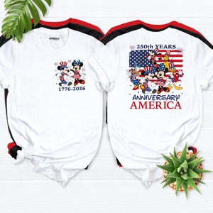 May include: Two white t-shirts with red and black trim. One shirt features Mickey and Minnie Mouse in patriotic attire with the text "1776-2026". The other shirt displays a US flag with Mickey, Minnie, and friends, and the text "250th YEARS ANNIVERSARY AMERICA".