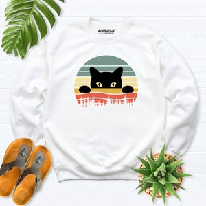 K&ouml;nnte beinhalten: Wei&szlig;er Pullover mit einem schwarzen Kater, der &uuml;ber einem Retro-Sonnenuntergang-Motiv lugt. Das Motiv ist ein Kreis mit einem Sonnenuntergang-Design in Orange-, Rot-, Gelb- und Gr&uuml;nt&ouml;nen. Die Katze ist schwarz mit gelben Augen und schaut &uuml;ber den oberen Rand des Kreises.