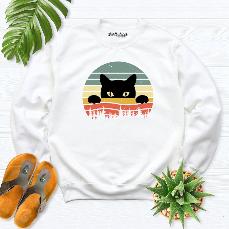 K&ouml;nnte beinhalten: Wei&szlig;er Pullover mit einem schwarzen Kater, der &uuml;ber einem Retro-Sonnenuntergang-Motiv lugt. Das Motiv ist ein Kreis mit einem Sonnenuntergang-Design in Orange-, Rot-, Gelb- und Gr&uuml;nt&ouml;nen. Die Katze ist schwarz mit gelben Augen und schaut &uuml;ber den oberen Rand des Kreises.