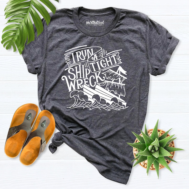 May include: A dark gray t-shirt with a white graphic design that says "I run a ship tight wreck". The graphic design features a ship with a sail and waves.