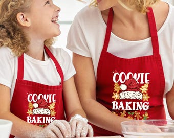 May include: Two red aprons featuring a white graphic print that reads COOKIE BAKING, accompanied by an illustration of a Santa hat surrounded by gingerbread cookies and stars. The aprons have neck loops and waist ties, designed for matching sets.