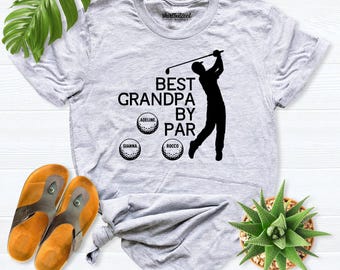 May include: A light gray short-sleeve t-shirt featuring a black graphic of a golfer and the text BEST GRANDPA BY PAR. Three golf ball illustrations are included, each labeled with the names Adeline, Gianna, and Rocco in black text.