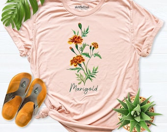 Op de afbeelding: Een lichtroze T-shirt met korte mouwen en een botanische illustratie van oranje en gele goudsbloemen met groene bladeren en stengels. De tekst "Marigold" is in het donkergroen onder de bloemen gedrukt en "shirtbutcool" staat bij de hals.