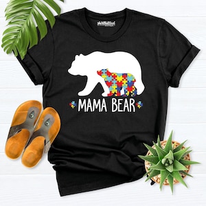 Pode incluir: Uma t-shirt preta com a silhueta branca de um urso grande que contém um urso mais pequeno. O urso mais pequeno está preenchido com um padrão de peças de puzzle coloridas. Abaixo dos ursos, o texto MAMA BEAR está escrito a branco, ladeado por dois pequenos ícones de peças de puzzle.