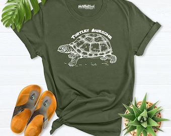 May include: An olive green short-sleeve t-shirt featuring a white line-art illustration of a turtle in the center. Above the turtle, the text reads "TURTLEY AWESOME" in a white arched font. The shirt has a crew neckline and a brand label.