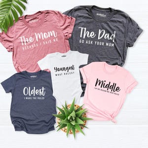 Pode incluir: Uma coleção de cinco camisas e um body de bebé com texto impresso. A camisa rosa diz The Mom BECAUSE I SAID NO, a cinzento-escura The Dad GO ASK YOUR MOM, o body branco YOUNGEST WHAT RULES, a azul-marinho OLDEST I MAKE THE RULES e a rosa-clara MIDDLE I'M THE REASON WE HAVE RULES.