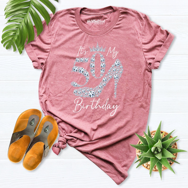 May include: A mauve t-shirt featuring the text "It's My 50th Birthday" and a high-heeled shoe design made of sparkling gems. The shirt is folded, with brown sandals and a small succulent plant visible.