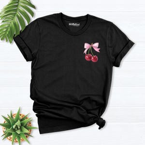 May include: Black t-shirt with a pink bow and two red cherries on the front. The shirt has a crew neck and short sleeves.