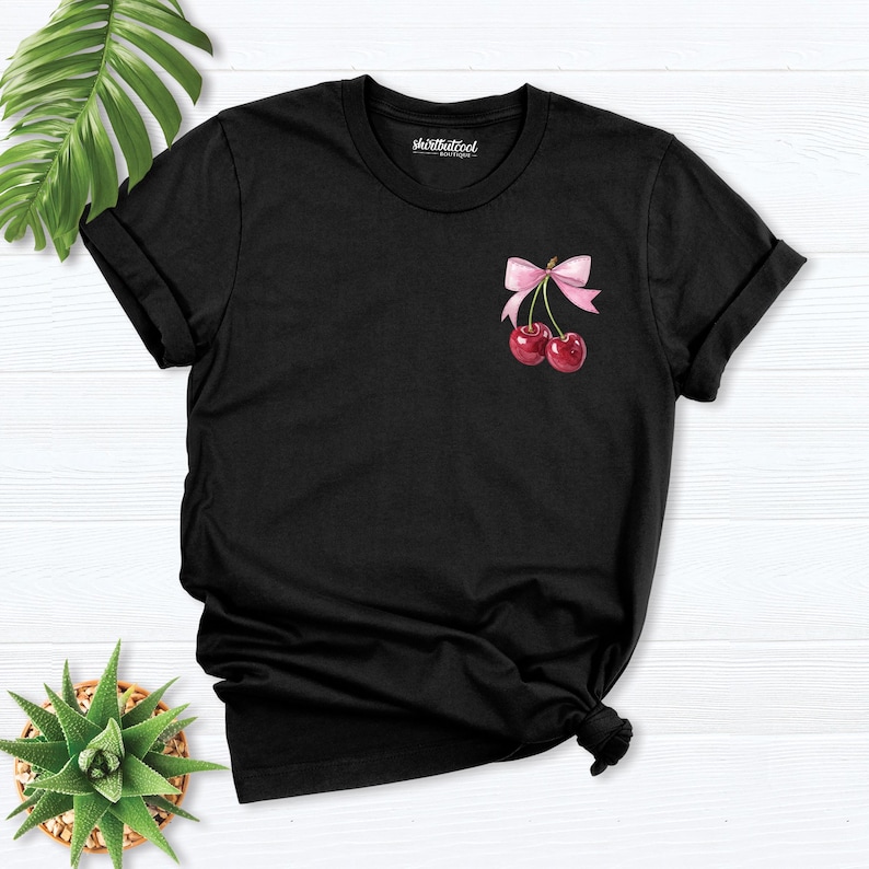 May include: Black t-shirt with a pink bow and two red cherries on the front. The shirt has a crew neck and short sleeves.