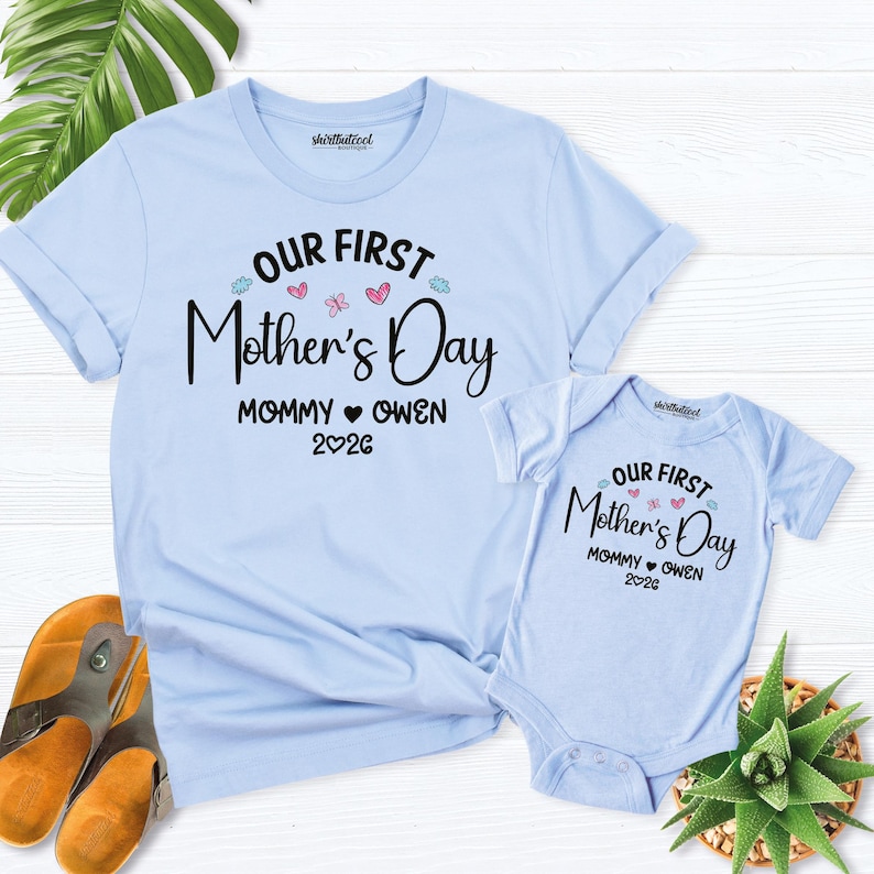 May include: Light blue t-shirt and baby onesie set with the text "OUR FIRST Mother's Day" in black script. The text "MOMMY OWEN 2026" is below. The set includes a short-sleeved t-shirt and a short-sleeved onesie.