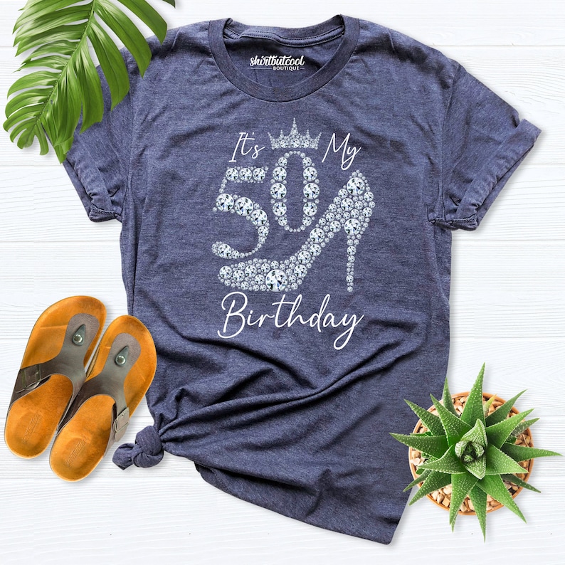 May include: A navy blue t-shirt featuring the text "It's My 50th Birthday". The number 50 is formed with sparkling gems, and the zero is a high heel. The shirt is a casual style, perfect for a milestone celebration.